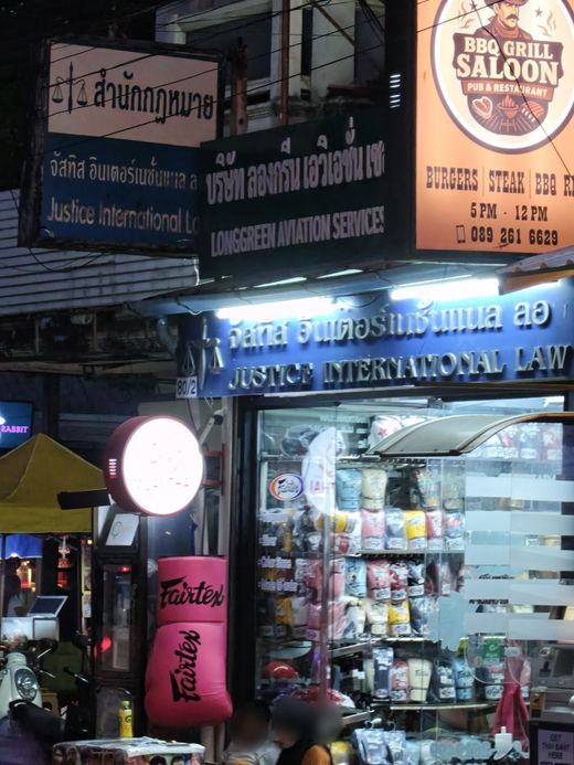 Signs stacked at 80/2 Loi Kroh: Justice International Law, Longgreen Aviation Services, BBQ Grill Saloon, a Fairtex bag outside the fight-gear shop