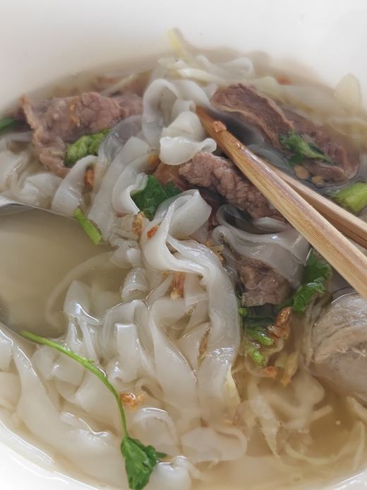 A bowl of wide rice noodles with beef, chopsticks lifting a strand