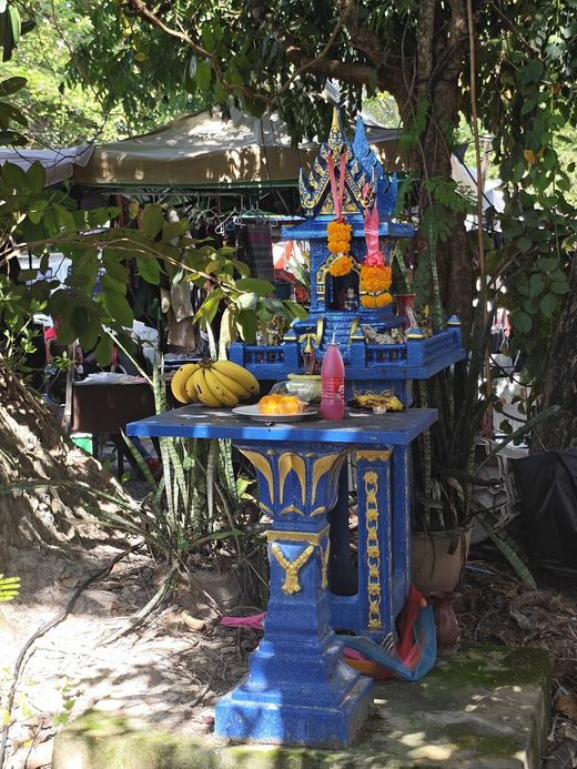 A blue spirit house with bananas and red drinks at the edge of the market