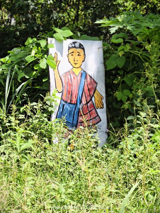 A painted cut-out boy with a shoulder bag waves from the grass