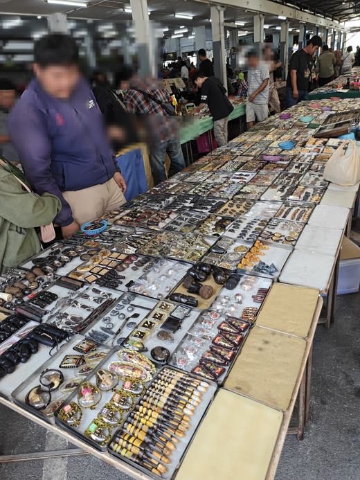 A long table of amulet trays in the market hall, buyers along it