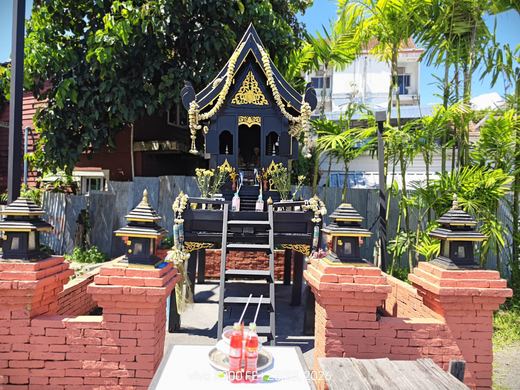 A black-and-gold spirit house on a brick base with red drinks, small shrines either side