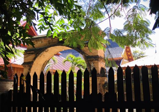 A brick arch and a Lanna roof seen over a dark picket fence