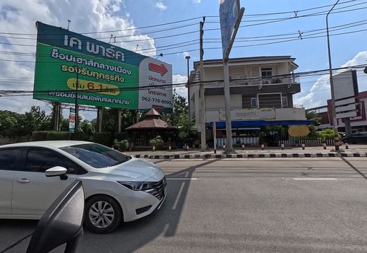 A big green billboard for the K Park shophouse project with a teak pavilion, and a three-storey building with the Machita sign; a white car alongside.