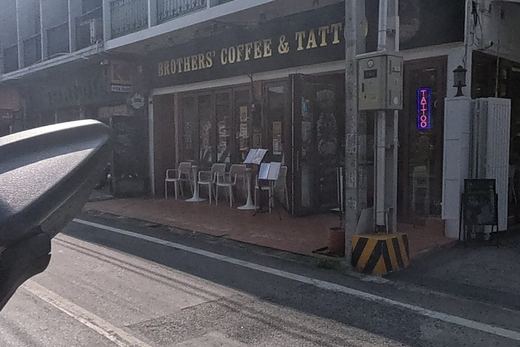 A corner cafe and tattoo shop with outdoor seating sits beside a driveway with a small white spirit house on its wall.