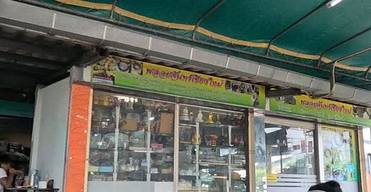 The พลอยจันทร์เชียงใหม่ shopfront with its glass cabinets, a table of amulet trays in front, and a seller smiling at the camera.