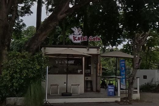 A small free-standing kiosk cafe, Katai Cafe, with outdoor seating under umbrellas beside a noodle shop sign advertising Coca-Cola.