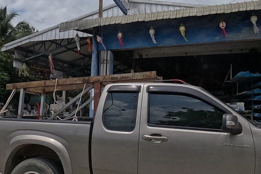 A pickup truck in front of a blue-fronted building-materials shop with keys and tools hanging over the open doorway, a small gold spirit house on a pedestal fur