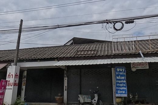 A row of shophouses: a metalwork/repair shop with a blue sign and phone number, a gel-nail shop's pink sign, and a small restaurant with potted plants and a woo