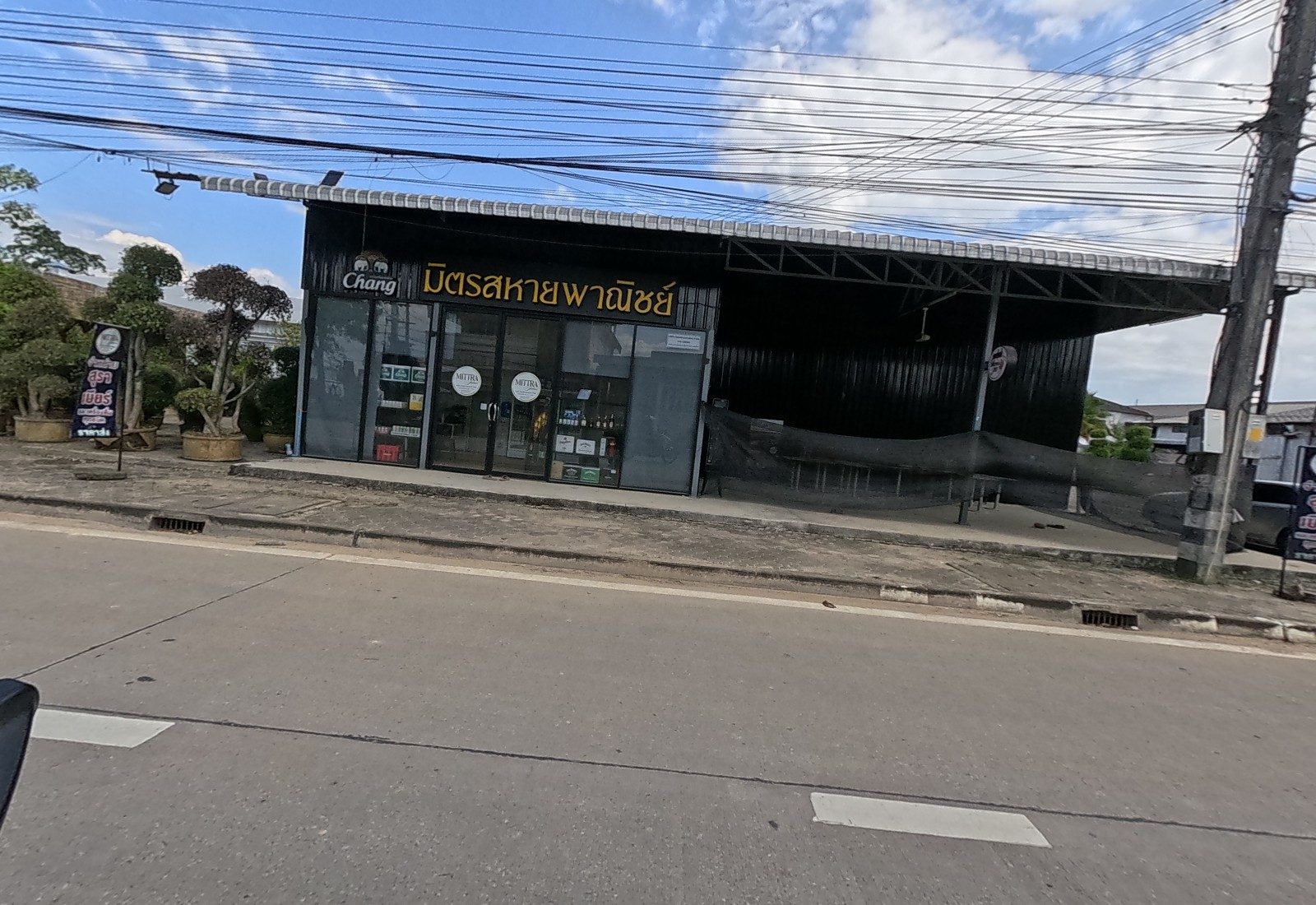 A black steel shop signed มิตรสหายพาณิชย์ with a Chang logo and glass doors, a bonsai tree display beside it and a standing beer sign.