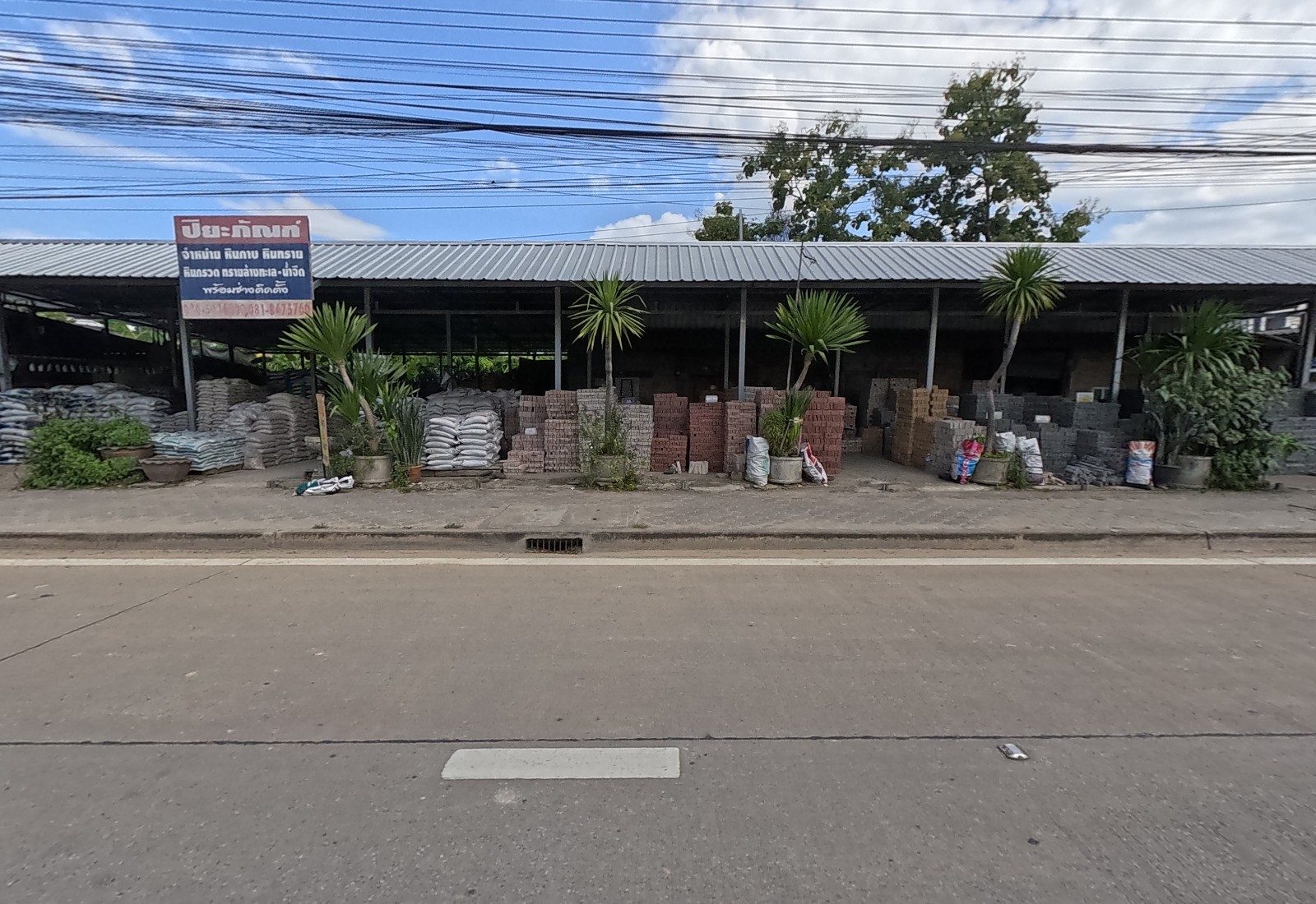 A long open shed of sacks, bricks and paving blocks behind potted palms, under the red-and-blue ปิยะภัณฑ์ sign.