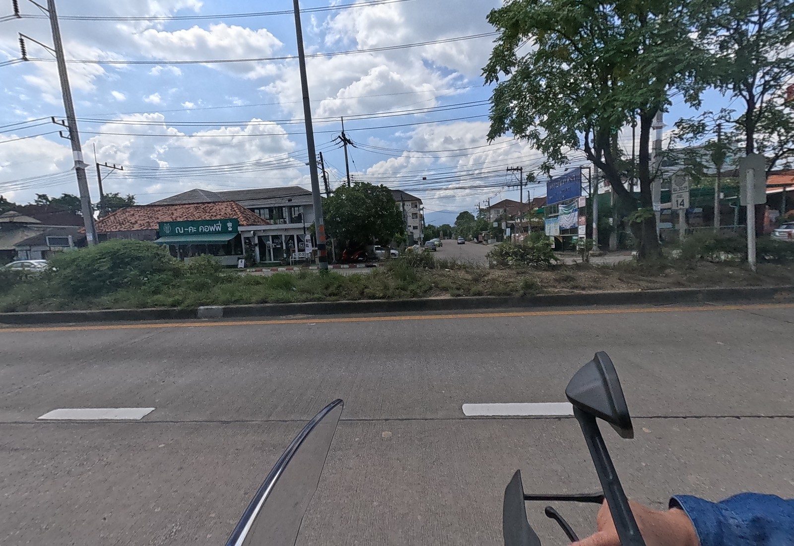 Across the median, the green ณ-คะ คอฟฟี่ café under a tiled roof at the corner of a side road, a blue sign board and a white road marker with 14 on it.