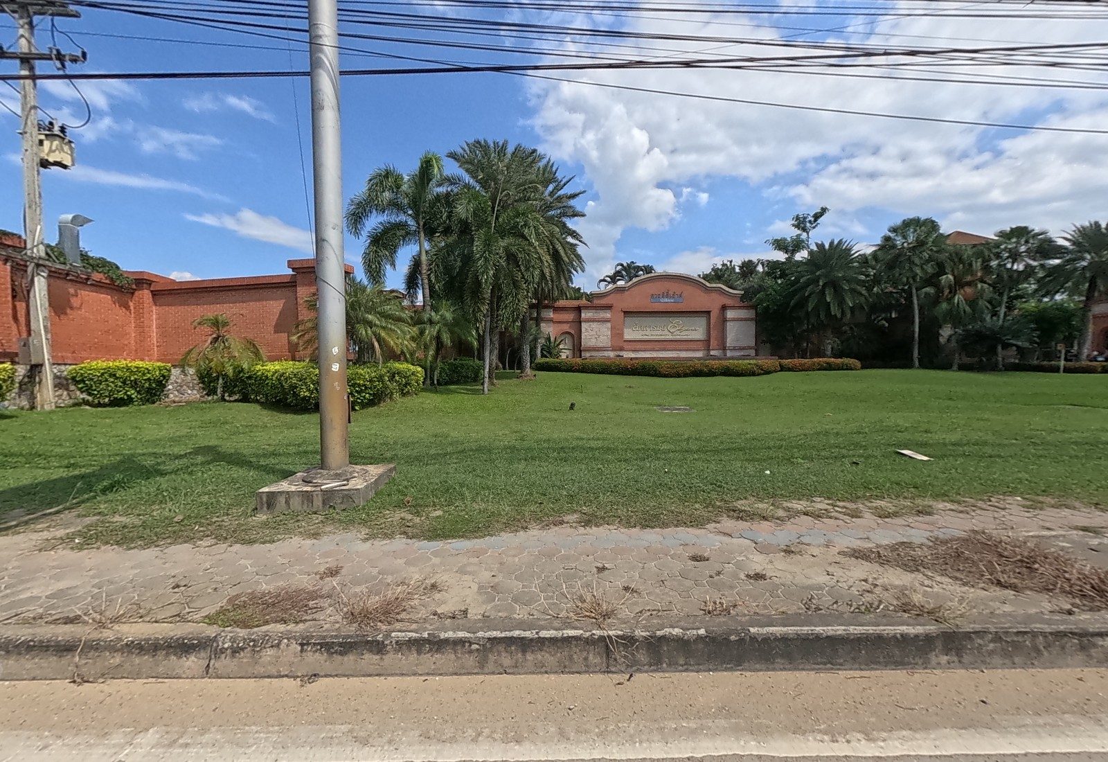 A lawn with palm trees in front of a terracotta-walled housing estate entrance signed ลัดดารมย์ Elegance.