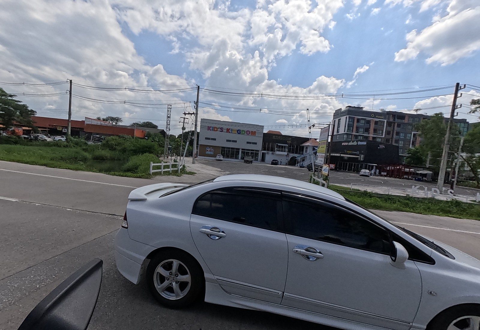 A white Honda Civic turns out beside the KID'S KINGDOM building and its big car park; white KID'S KINGDOM letters stand by the road and condo blocks rise behind.
