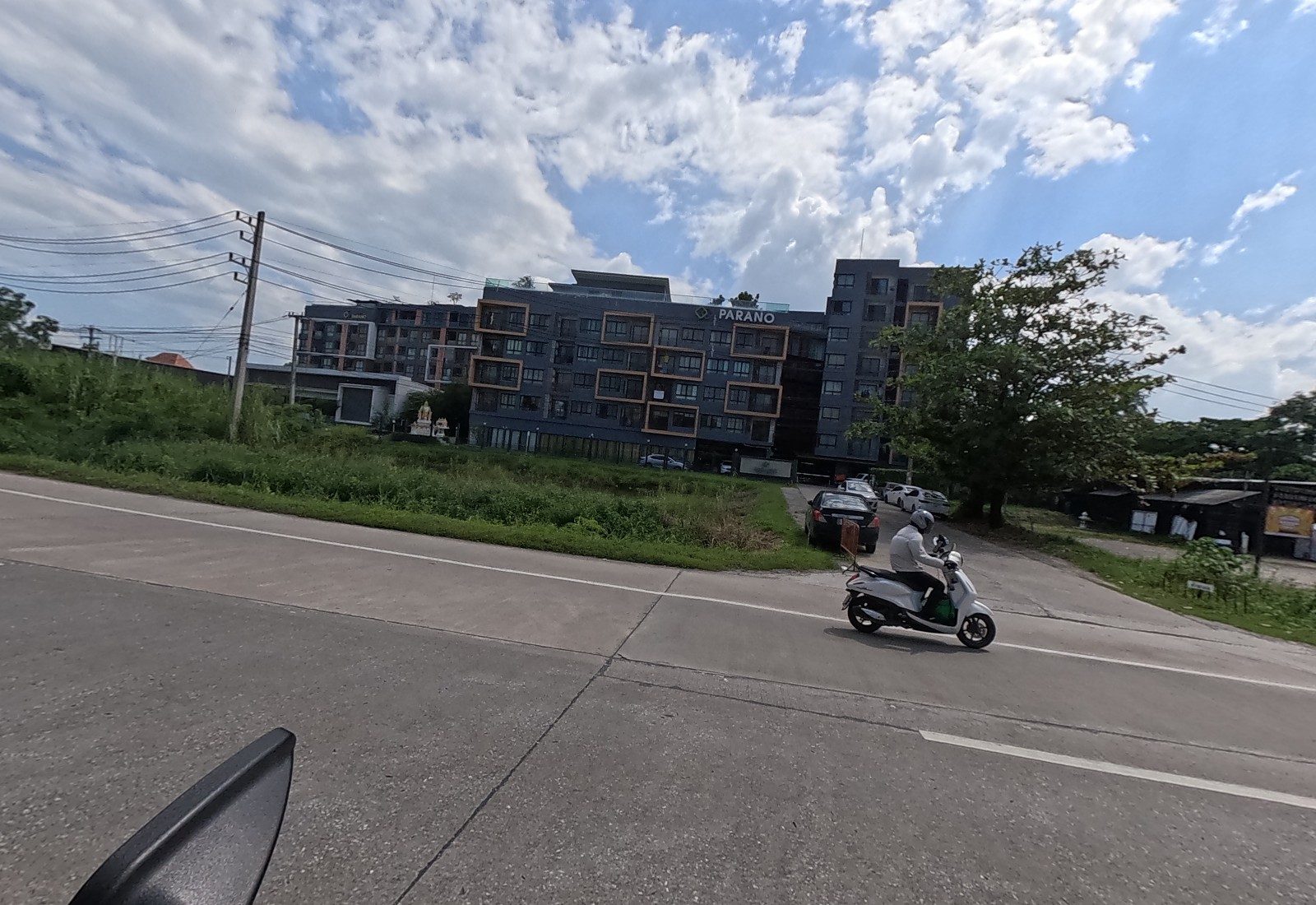 The PARANO condo stands behind a grassy lot; a man on a white scooter turns out of its drive, with a spirit house by the gate.