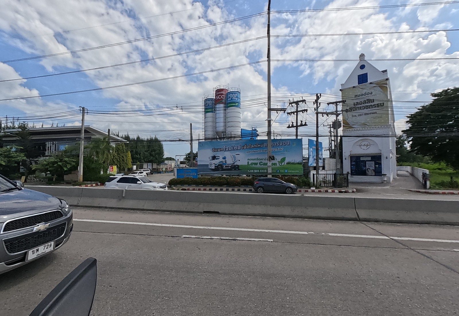 The CPAC concrete plant with its silos and 'Low Carbon' billboard, and the white gabled clinic 'หมออ๋อง เลอลลิน คลินิกเวชกรรม'; the grey Captiva alongside.