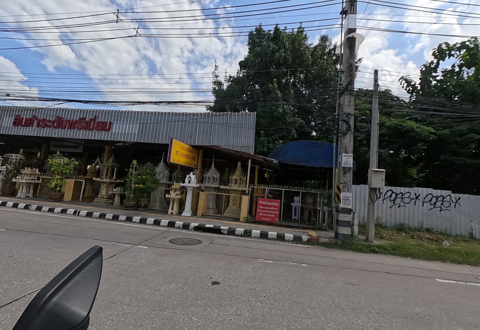 A spirit-house shop 'อ้อมเวียง ศาลพระภูมิ' displays rows of shrines under a roof reading 'สินค้าระดับพรีเมี่ยม'; a graffiti-tagged metal fence follows.