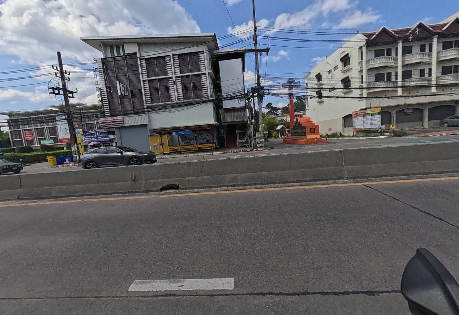 Across the road: a louvred building with a 7-Eleven, a yellow bus shelter, an orange monument at a side road and a community noticeboard.