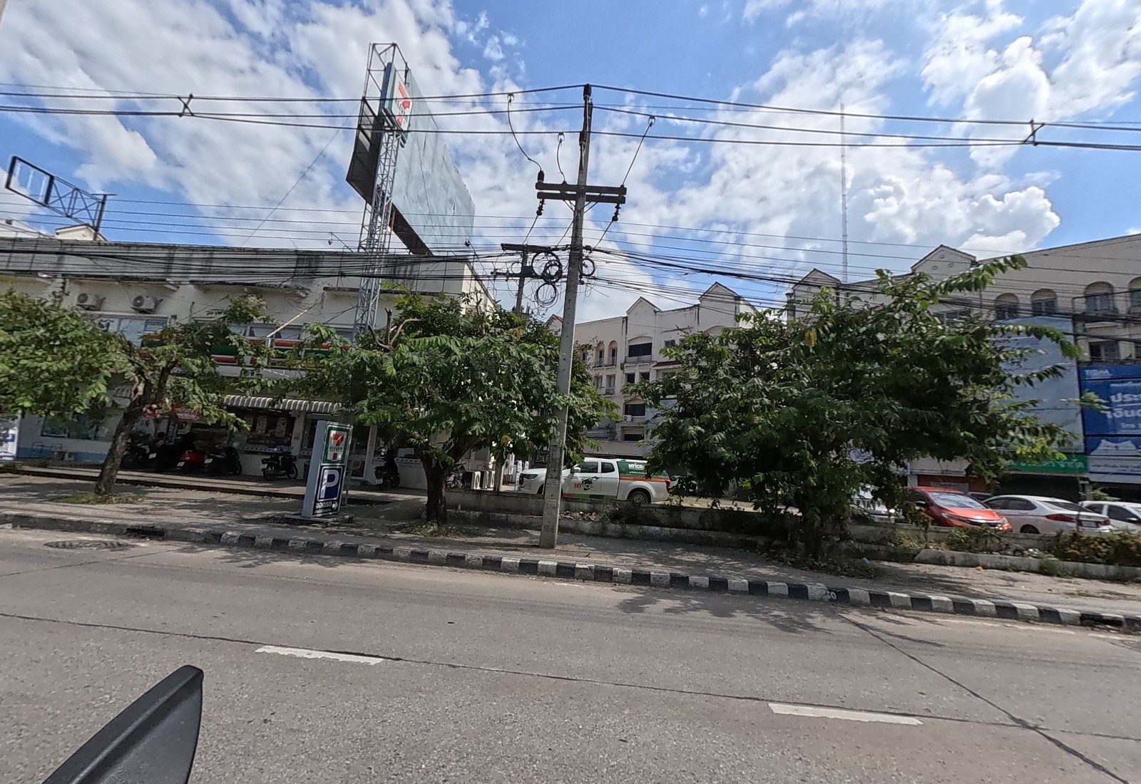 A 7-Eleven with a 7-ELEVEN/P pole sign in a low building, a big billboard frame above, and a four-storey row with a blue 'ประกัน เงินด่วน' loan sign.