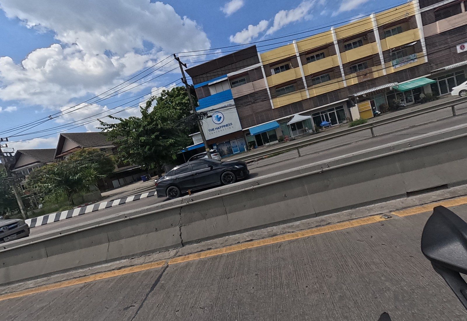 Across the barrier: THE HAPPINESS CORPORATION on a corner shophouse, a row of yellow four-storey shophouses, and a black Honda sedan.