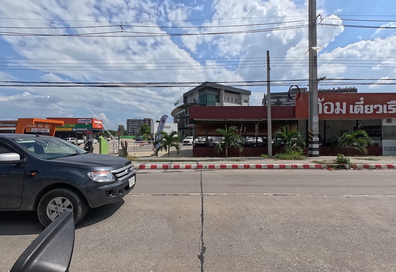 A red-fronted boat-noodle restaurant with open seating, a MINIMAL container café and a round office building behind, tyre and car-wash shops further left; a pickup at left.
