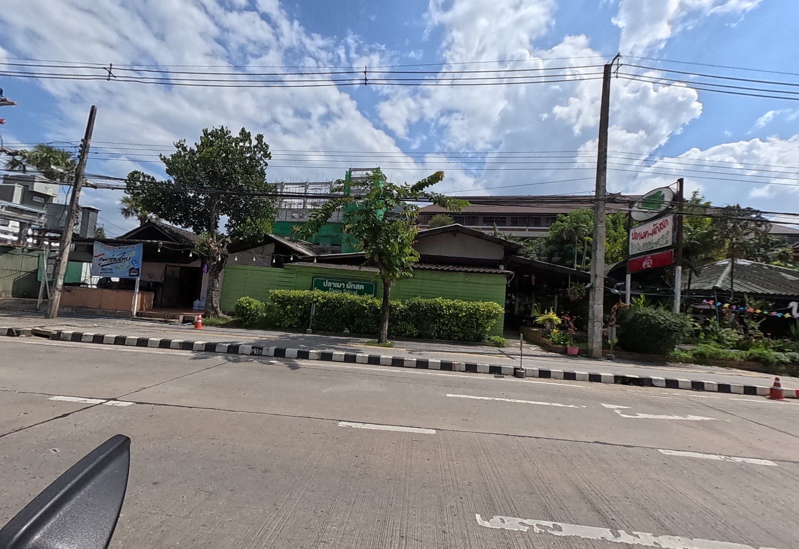 A green wooden garden restaurant with a green 'ปลาเผา ผักสด' sign and a pole sign, a drinks banner at a small shop on the left, the school's netted building behind.