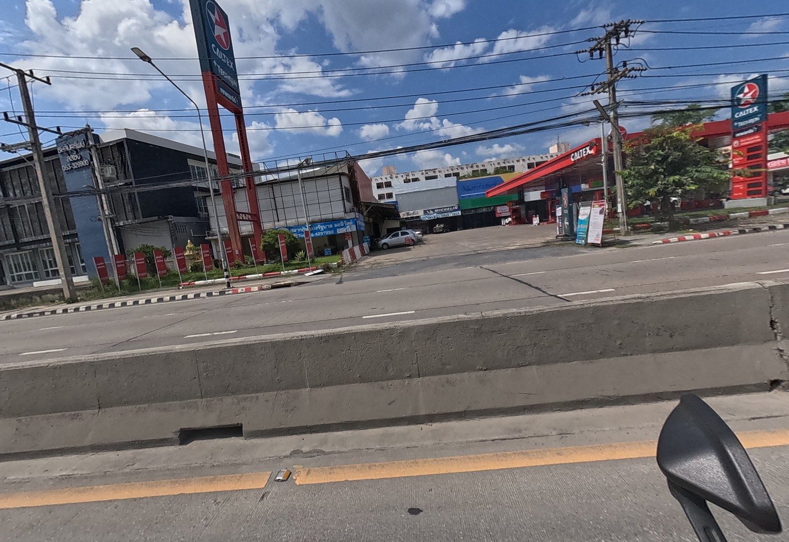 Across the road, the Caltex station with its tall pylon, a lottery-ticket shop with red flags, a Michelin tyre shop behind, and a blue-signed office building.