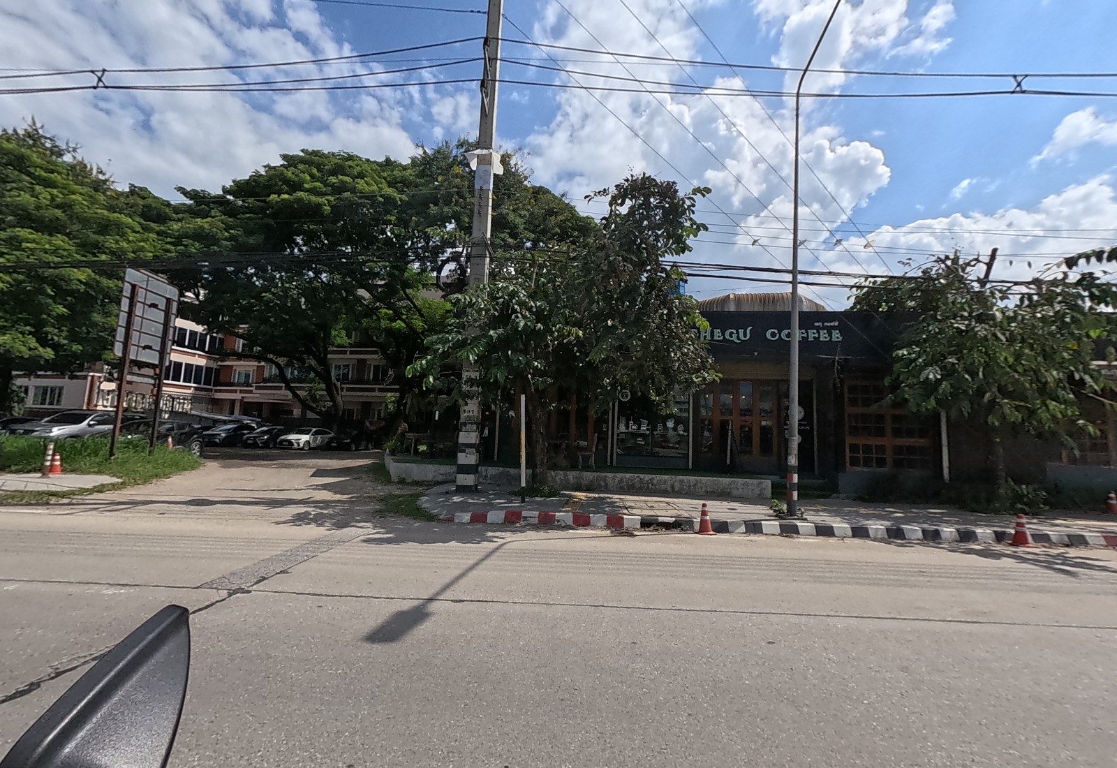 A dark wood-and-glass café with a CHEGU COFFEE fascia under trees, a side road and a car park in front of a pink apartment block to its left.