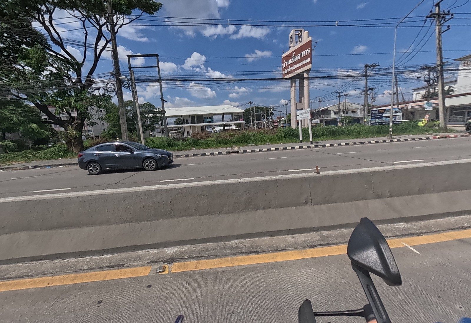 Across the median stands the tall red Baan Ua Arthorn sign beside a vacant grassy lot, with a small area-office direction sign and a billboard.
