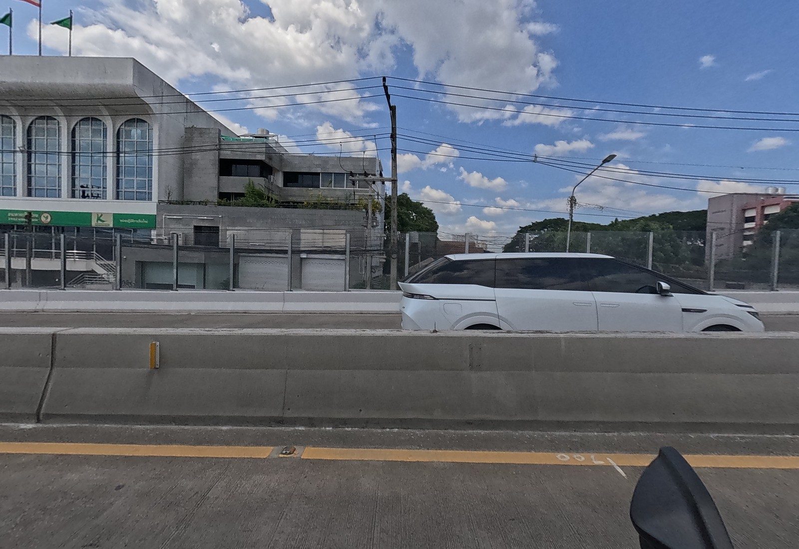 Across the flyover, a white Kasikorn Bank building with arched windows, a for-rent/for-sale billboard and an MPV passing.