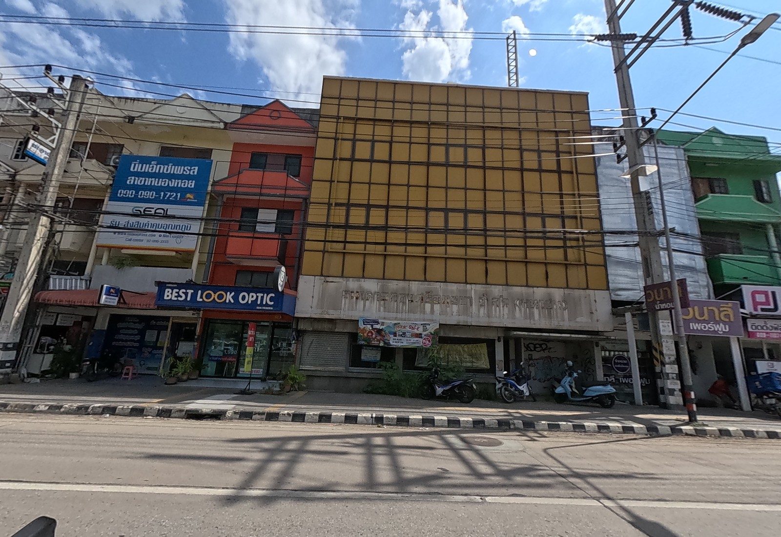 A shophouse row: a parcel-delivery branch sign, Best Look Optic, a derelict yellow-panelled building with a book-sale banner, a perfume shop and PJ Autofilm.