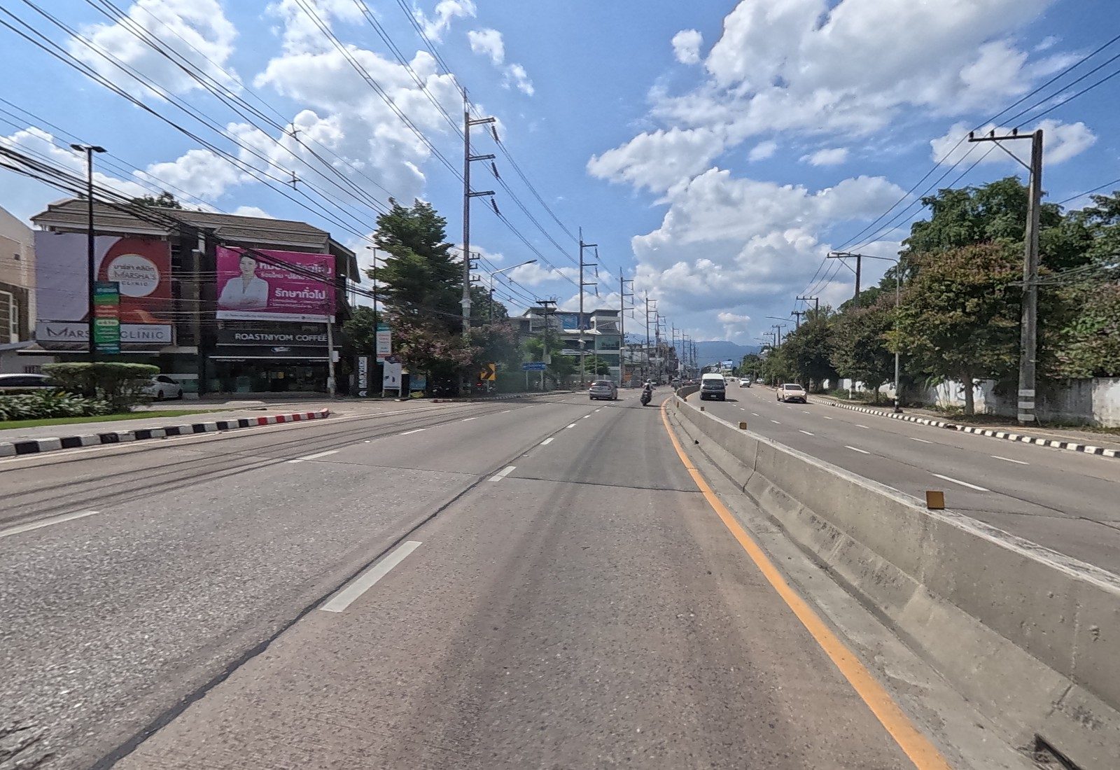 Mahidol Road ahead: Marsha's Clinic and Roastniyom Coffee on the left, a curve sign and blue signs further on, mountains on the horizon.