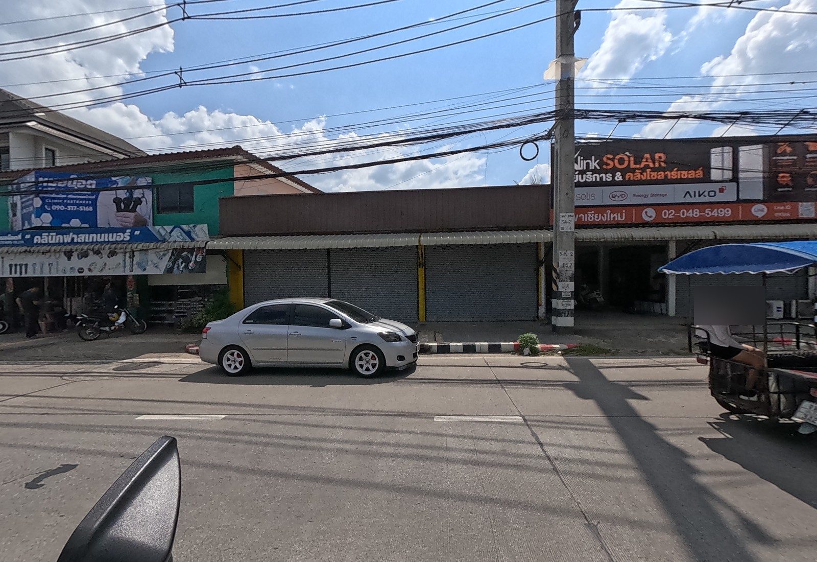 A fastener shop in a green building, a row of shuttered units, and a solar-panel warehouse with a brown fascia; a sidecar vendor passes.