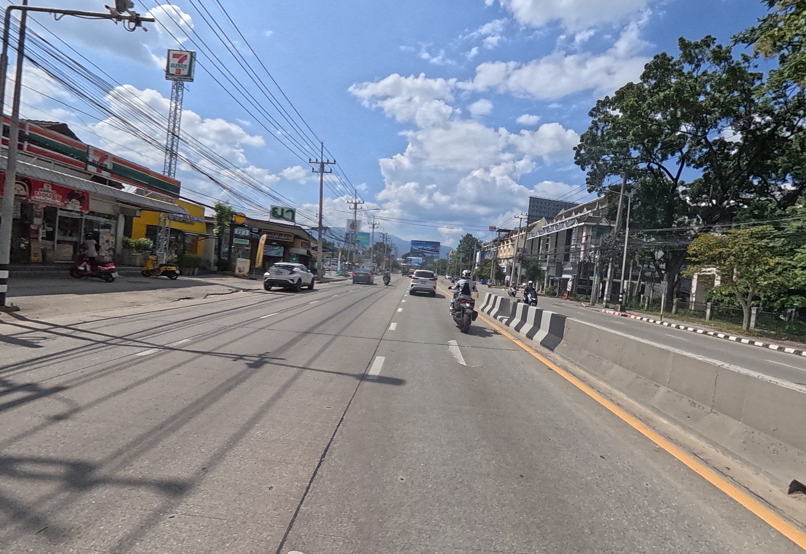 The 7-Eleven with its tall pole sign and a row of shops line the left kerb; a motorcyclist rides ahead.