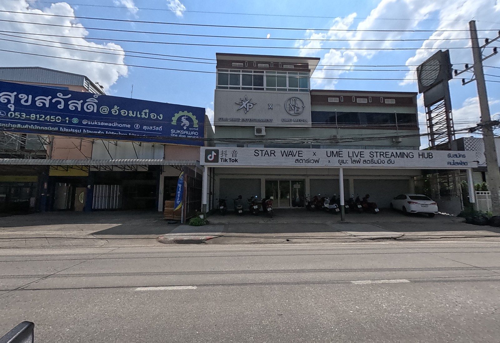 The Suksawad timber shop's long blue sign sits beside the Star Wave x UME live-streaming hub, with motorbikes parked in front.
