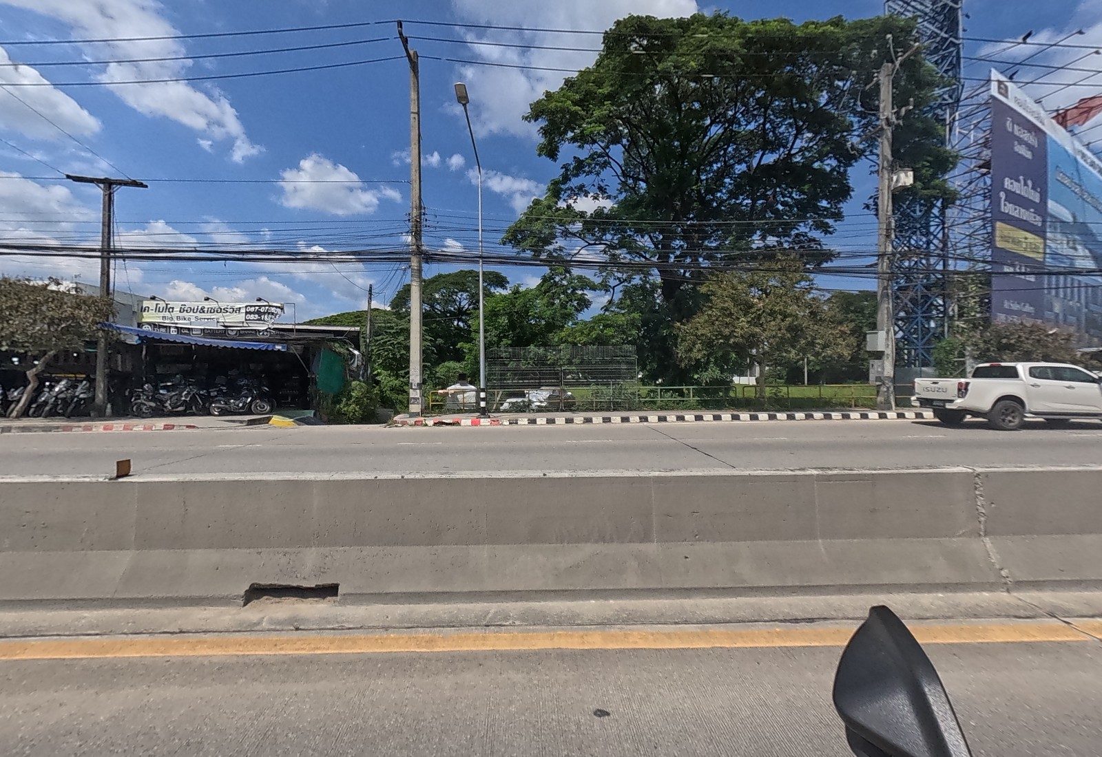 Across the median, big bikes are lined up under the K-Moto shop's blue awning beside a large tree and a condominium billboard.