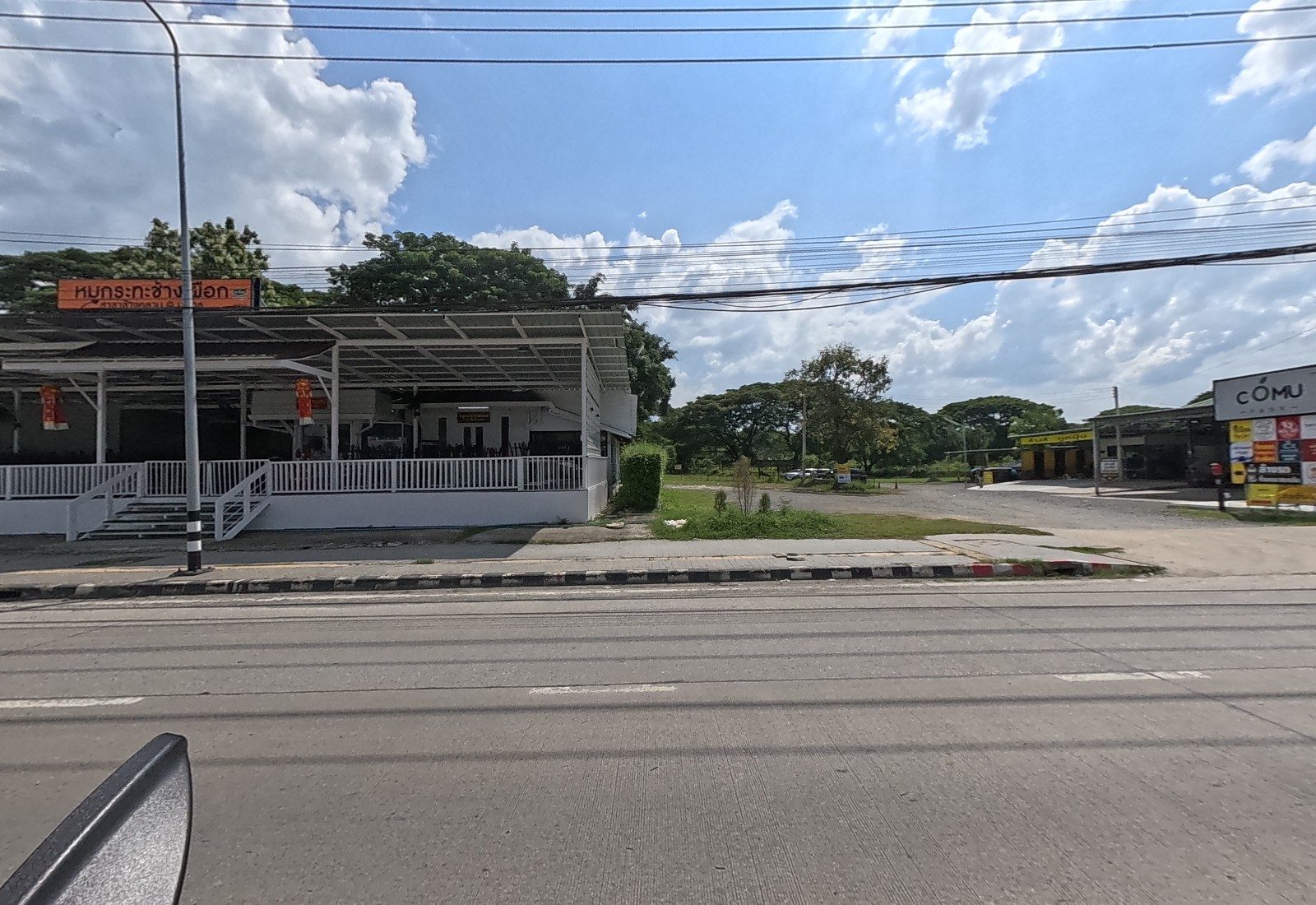 An open-sided moo kratha restaurant with a white railing sits under an orange sign, beside the entrance to the CÓMU plaza.