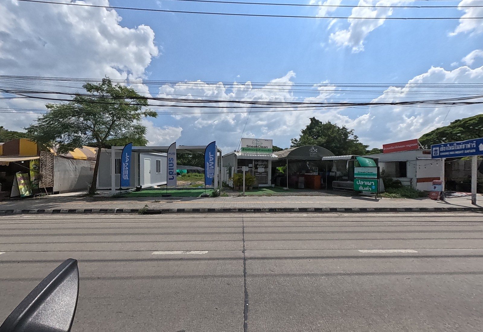 A row of small roadside businesses: a car-rental container office with JAMES flags, SneakClean shoe cleaning, a grilled-fish stall and a Chiang Mai Night Safari direction sign.