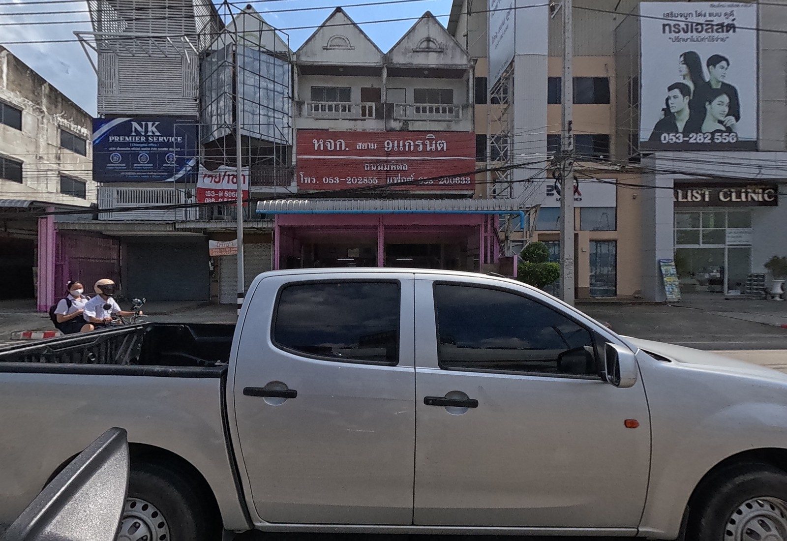 A row of shophouses: an NK Premier Service office, a granite shop with a red sign, and a clinic, with a pickup and two students on a motorbike in front.