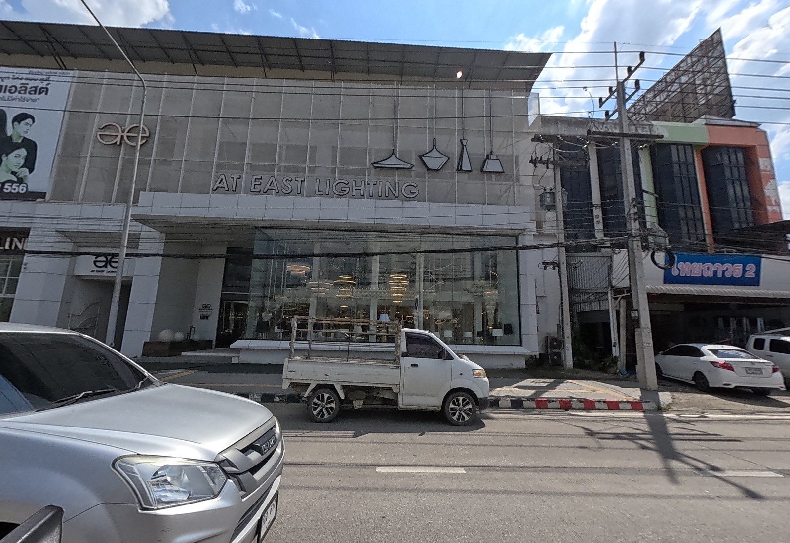 The AT East Lighting showroom, glass-fronted with pendant-lamp shapes on the facade, next to a shop signed ไทยถาวร 2.