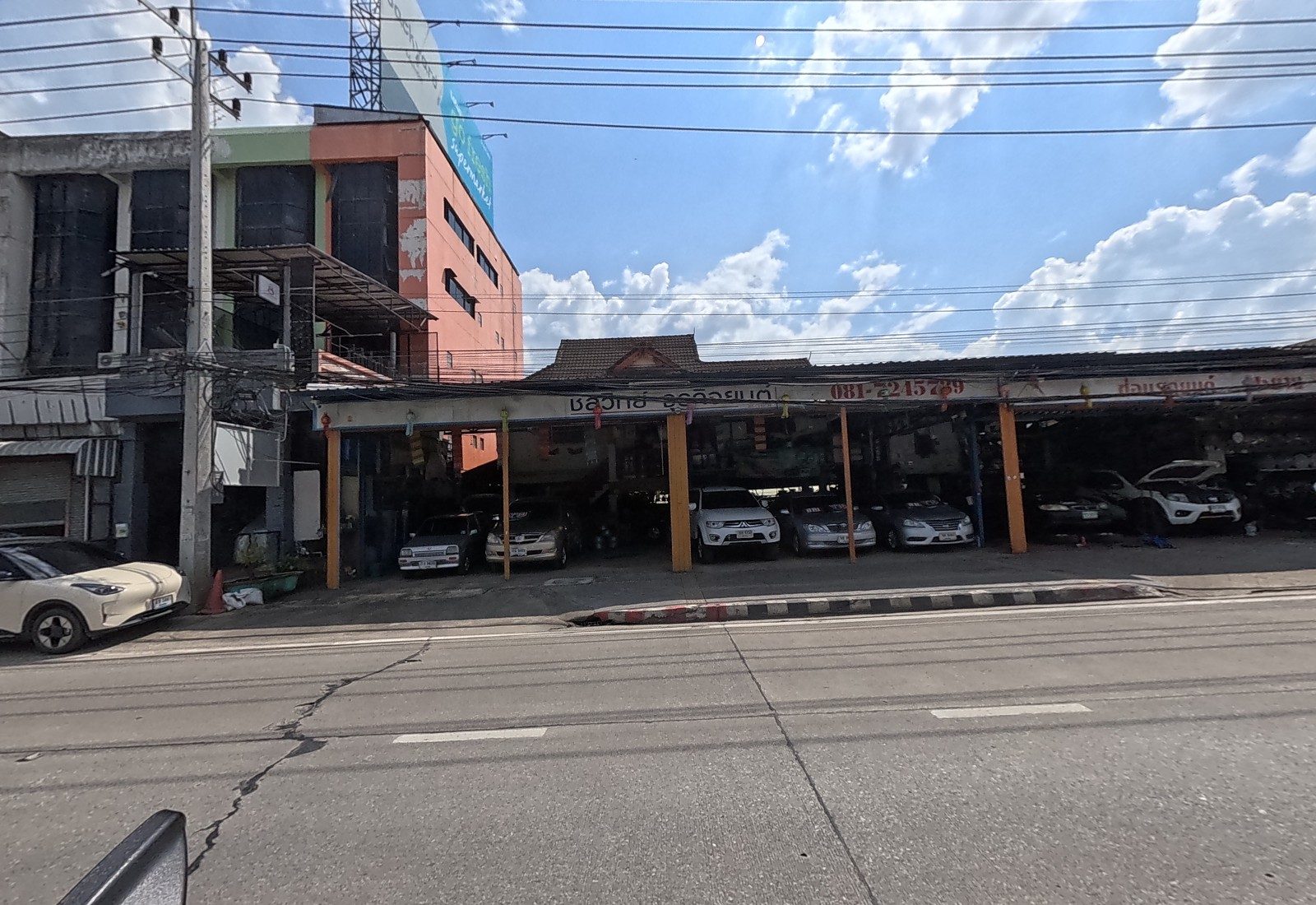 A long carport of used cars and a repair bay signed ชลวิทย์ ธุรกิจยนต์, beside an orange building with a supermarket billboard on top.