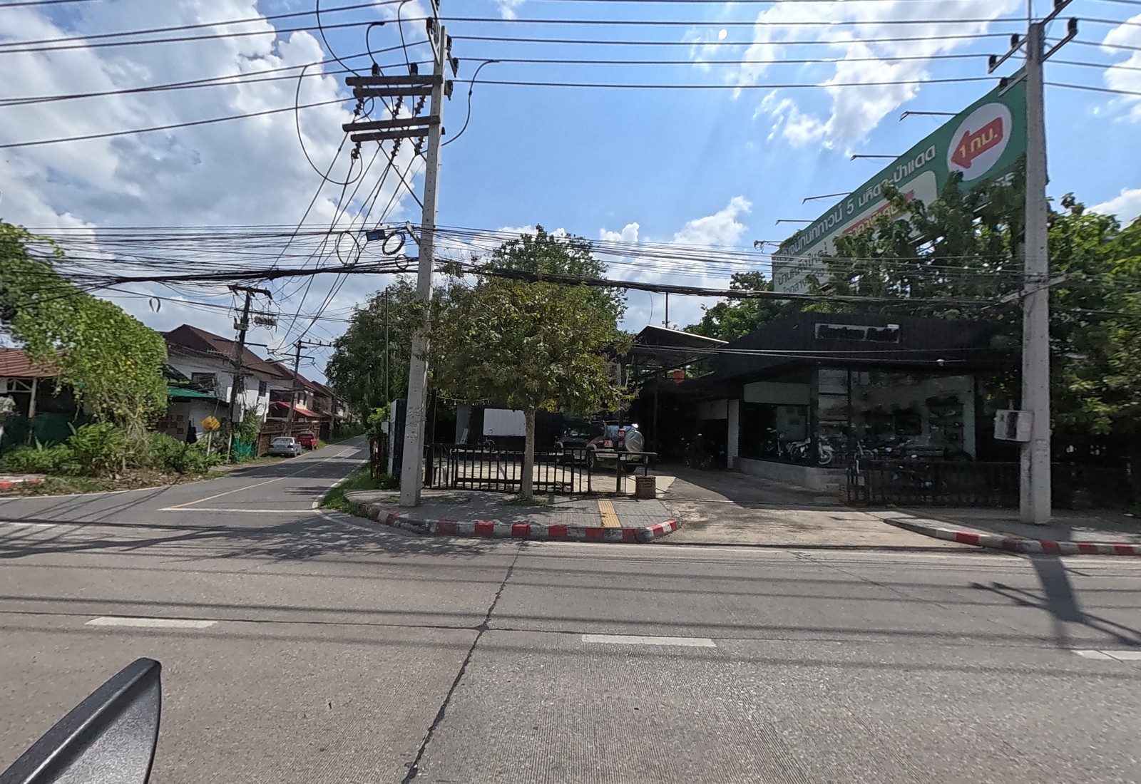 A black-fronted motorbike showroom on a corner with a side lane, under a big green billboard for a housing estate 1 km away.