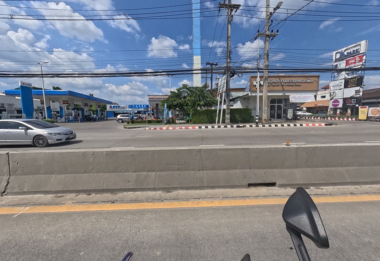 Across the road, the PTT station with a FIT Auto bay and a 7-Eleven, then the What the Pet vet clinic and the DK Park sign stack.