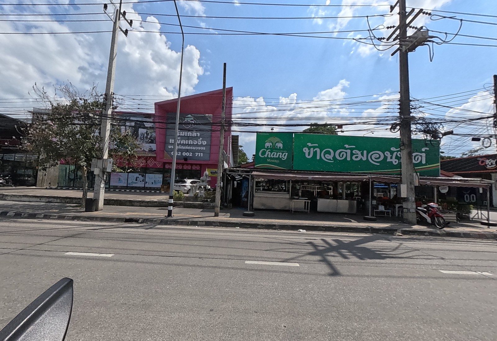 A pink building with a sign for Premium Glass (package deals) and a spirit house, beside the open-front Khao Tom Anan restaurant under a green Chang sign.