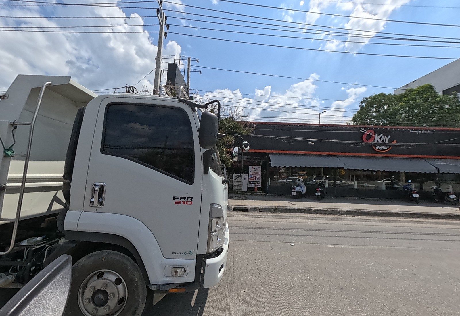 A white dump truck alongside hides most of the view; behind it, the long black-and-orange Okay Shabu restaurant with motorbikes parked.