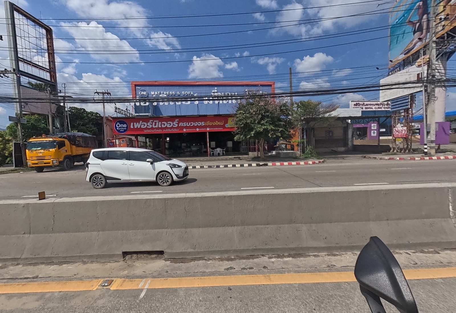Across the road, The One Furniture and Mattress & Sofa showroom under an orange-framed sign, a closed dental clinic building, and a PTT canopy at right.