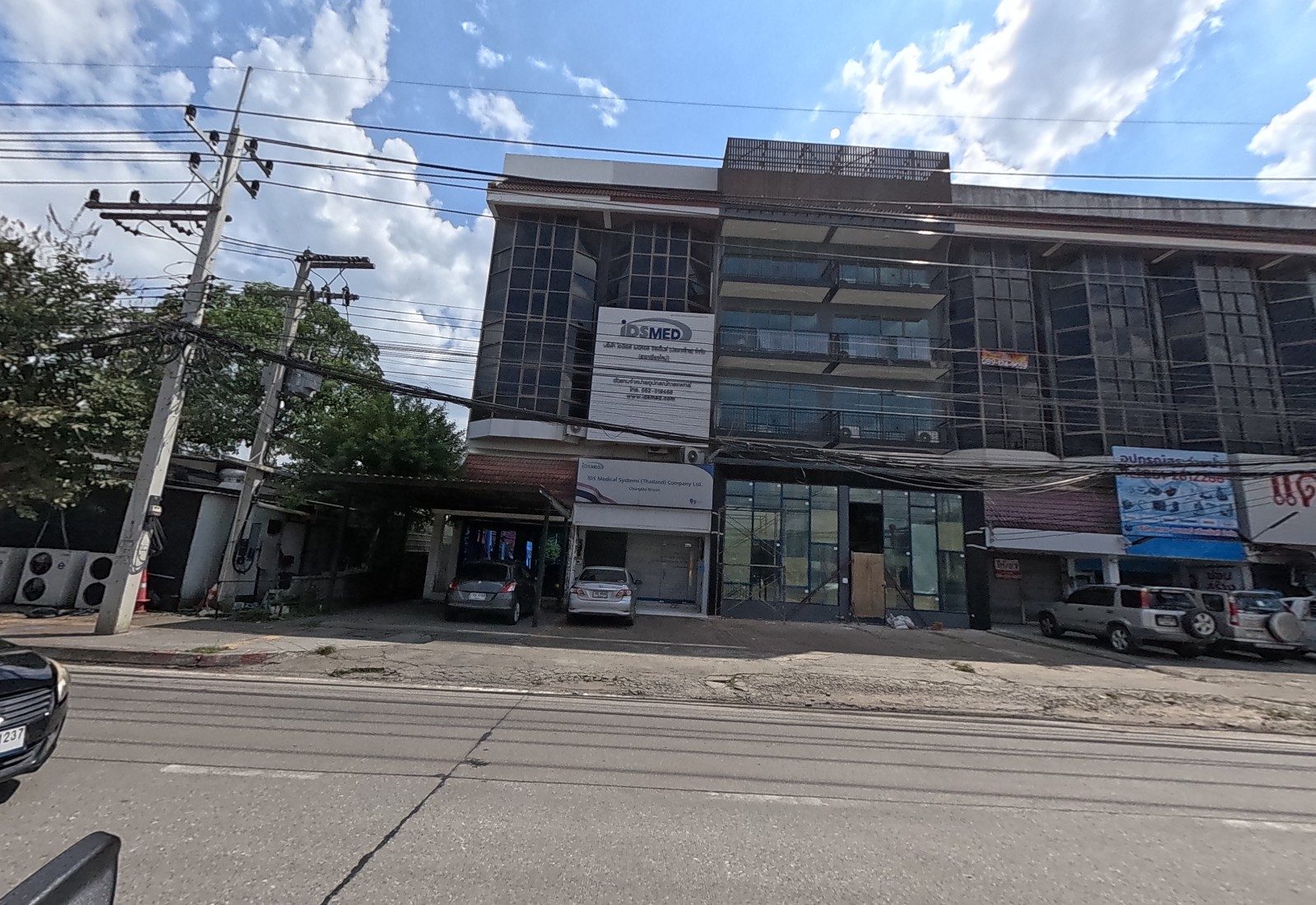 A dark glass office block housing IDS Medical Systems, beside a building being fitted out and a shop signed แดงกลอนประตู.