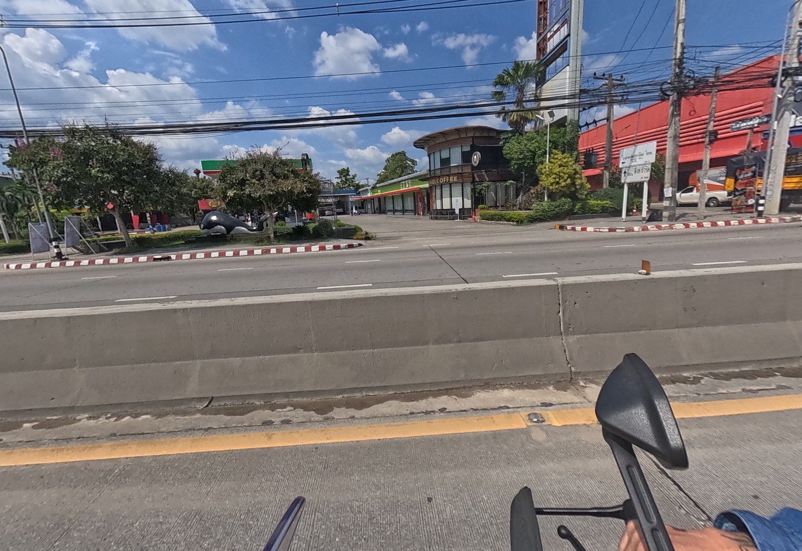 Across the road, a round café building signed …EL COFFEE with a killer-whale statue on the lawn, a petrol station canopy behind, and a red furniture store at right.