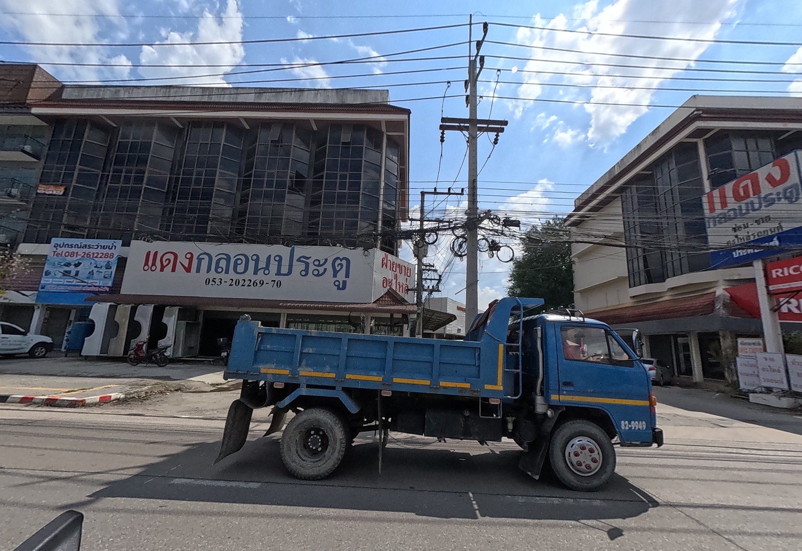 A blue dump truck passes the แดงกลอนประตู shop in a dark glass building, with a water-pump supplier's board, and a Ricoh office next door.