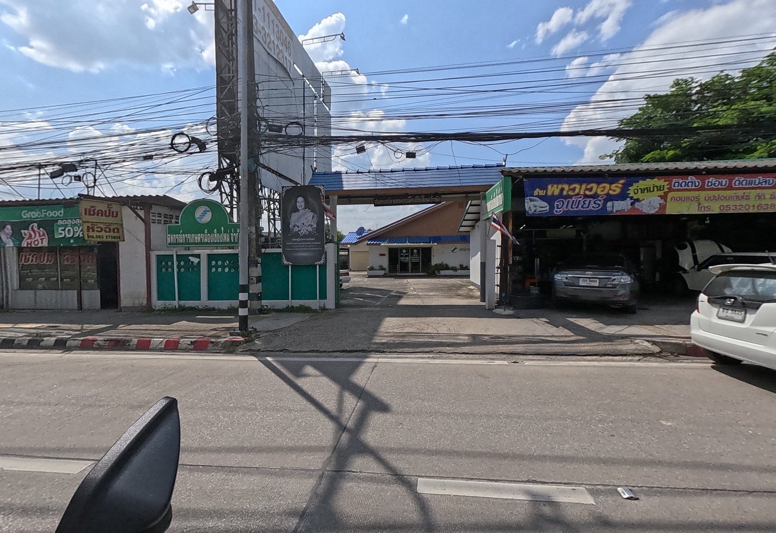 Gate of the Mueang Chiang Mai Agricultural Cooperative with a memorial portrait, a nozzle-repair shop at left and the Power Junior car air-con garage at right.